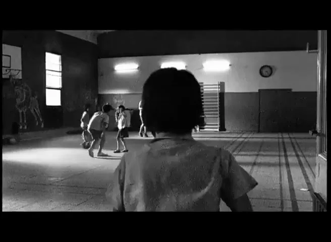 A child with short dark hair, seen from behind, runs towards other children playing in a dimly lit school gymnasium.