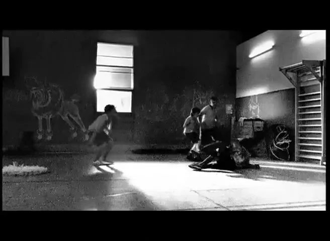 Four boys play in a dimly lit gym, with one boy on the floor and others running around him, in black and white.