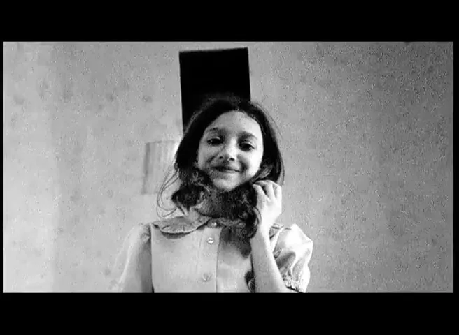 A young girl with long, dark hair, wearing a collared shirt, looks at the camera and smiles, in a black and white shot.