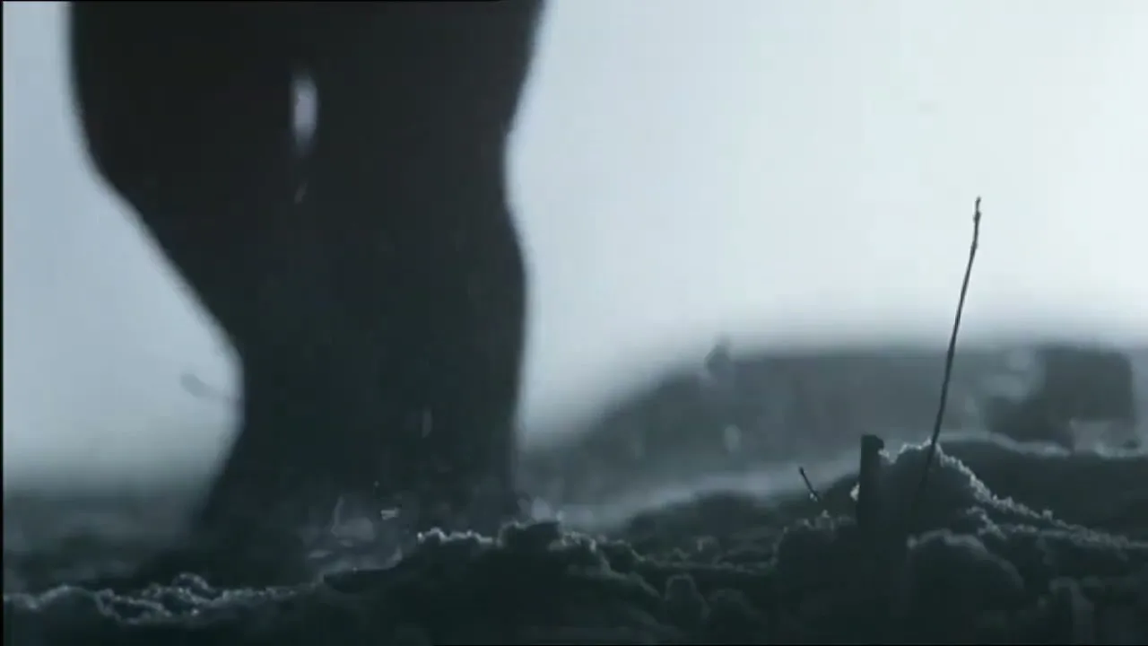 A person's bare feet walk on a dark, dusty surface, kicking up small particles, against a bright, hazy background.