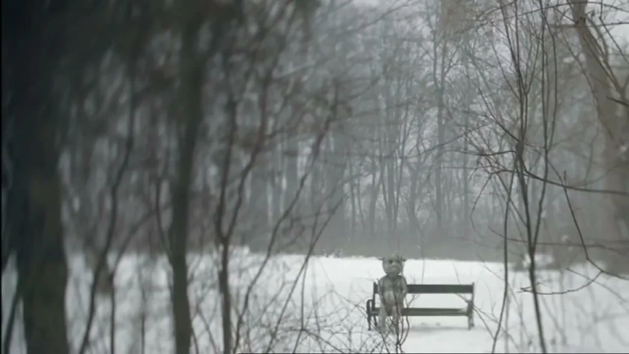 A small, grey, alien-like creature sits alone on a park bench in a snowy, desolate forest, looking sad.