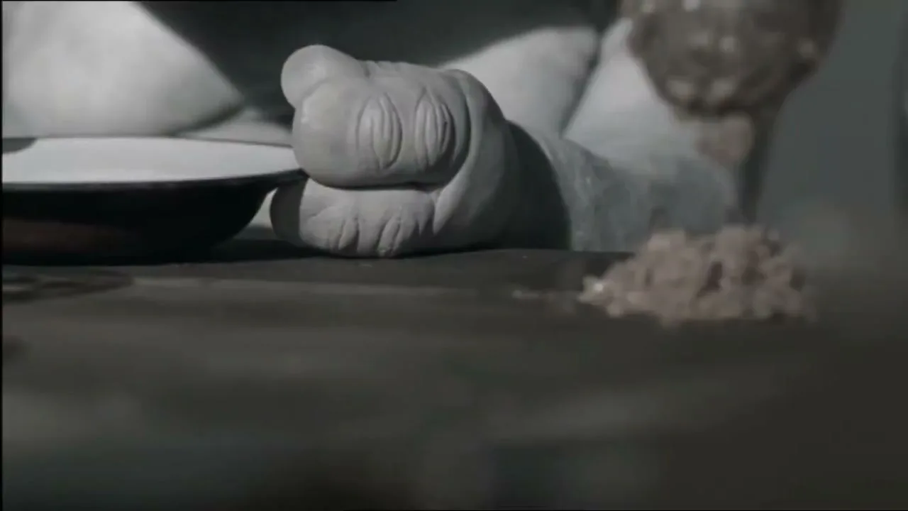 A person's foot, with wrinkled skin, is shown next to a small bowl on a dark surface, as a pile of dirt is dropped onto the surface.