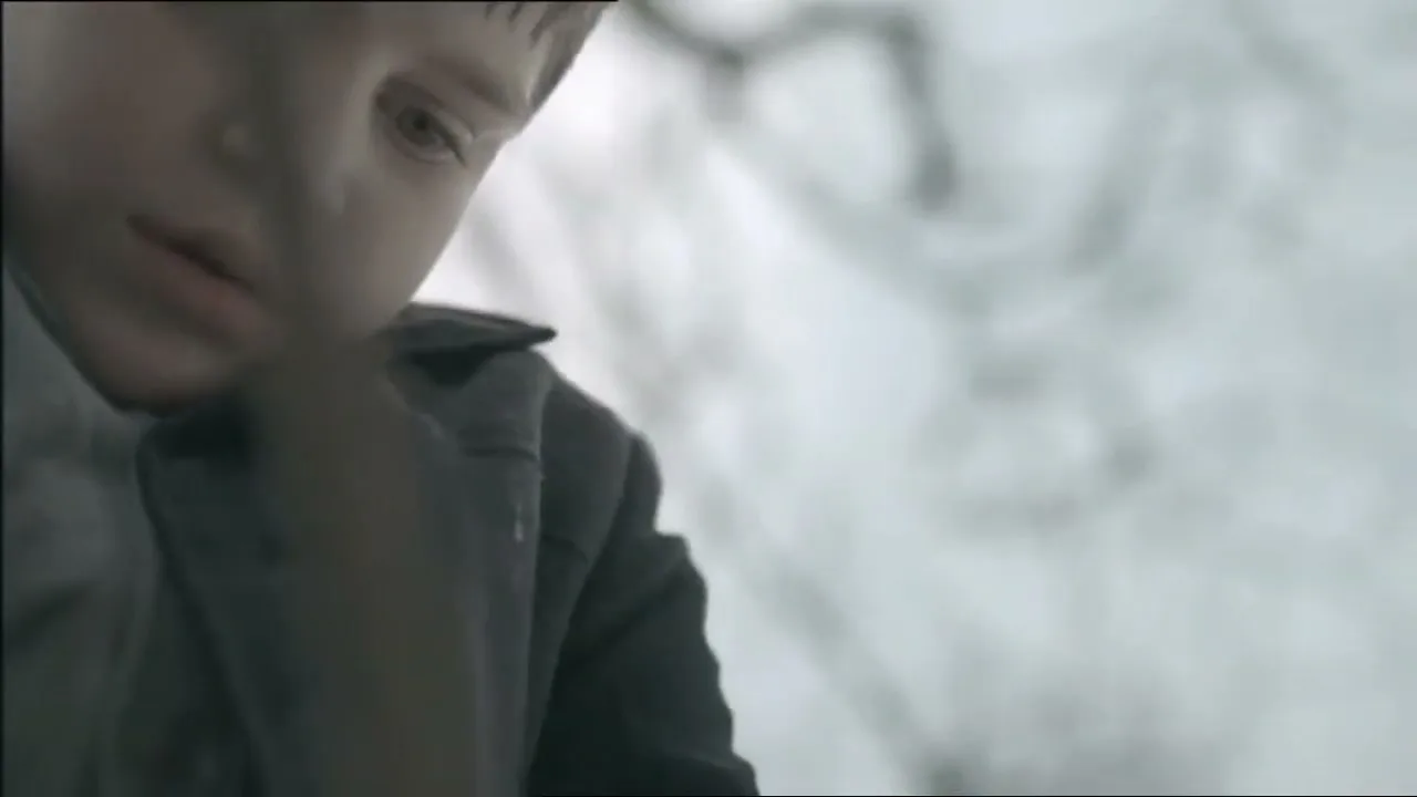 A young boy with short brown hair and a dark jacket looks down with a neutral expression in a snowy, desolate landscape.