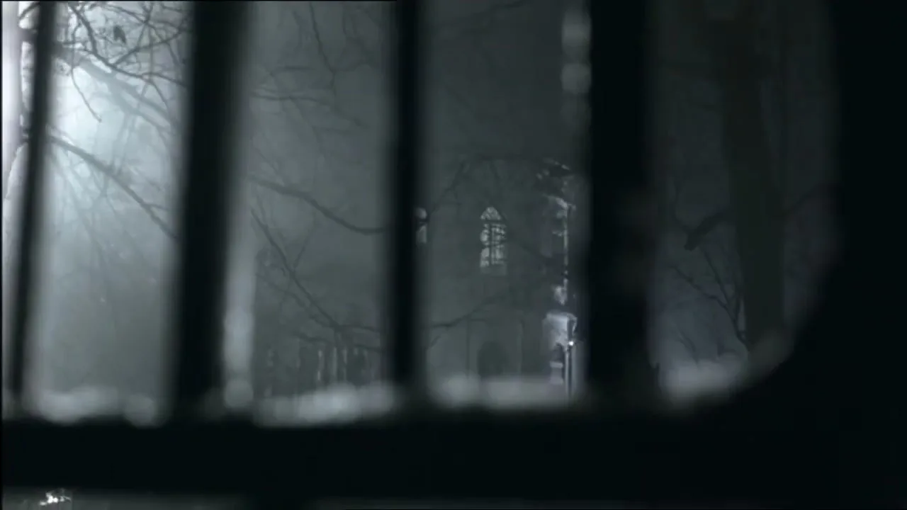 A blurry shot through prison bars reveals a spooky, fog-laden building with bare trees in the background, creating an eerie atmosphere.