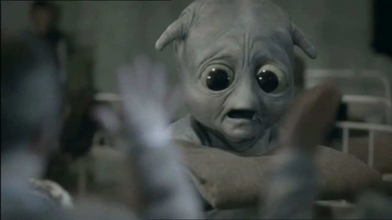 A small, grey alien with large, dark eyes looks sad while being held by a person, with other people in the background.