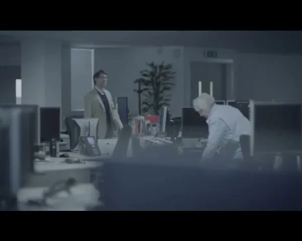 Two men, one younger and one older, are in an office, with the older man bending over his desk while the younger man stands nearby.