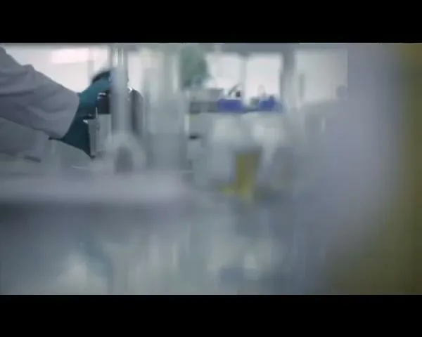 A person in a lab coat and blue gloves works with beakers and scientific equipment in a brightly lit laboratory.