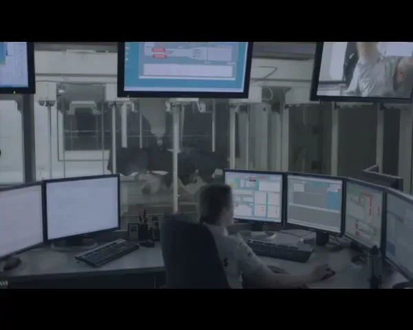 A female technician sits at a desk surrounded by multiple computer monitors displaying data in a control room.