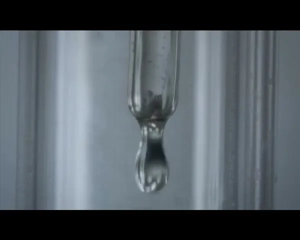 A clear liquid droplet forms and falls from a glass tube in a laboratory setting, highlighting precision and scientific process.