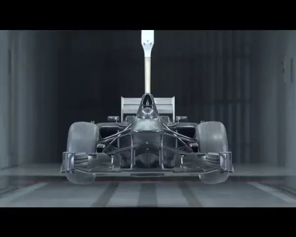 A dark, unpainted Formula 1 race car chassis is suspended in a sterile, industrial spray booth, ready for painting.