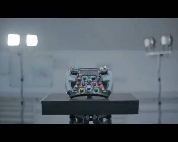 A Formula 1 steering wheel is displayed on a black pedestal in a brightly lit, sterile studio environment.