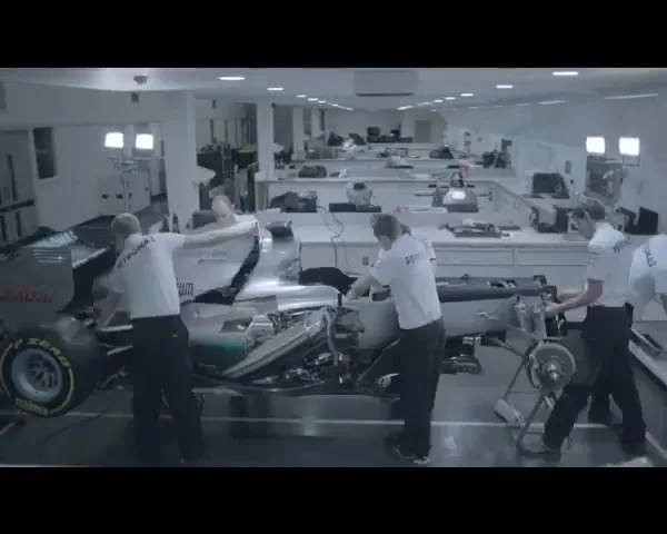 A team of mechanics in white shirts and dark pants work on a silver and green Formula 1 race car in a brightly lit garage.