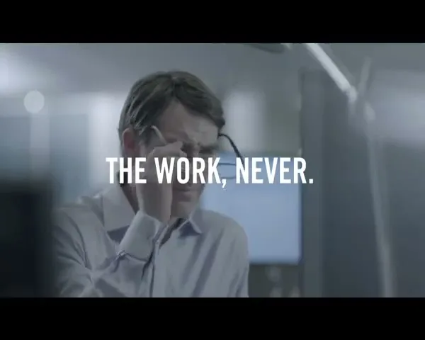 A man in his 40s, wearing a light blue shirt, puts on his glasses while looking intently at a computer screen in a modern office.