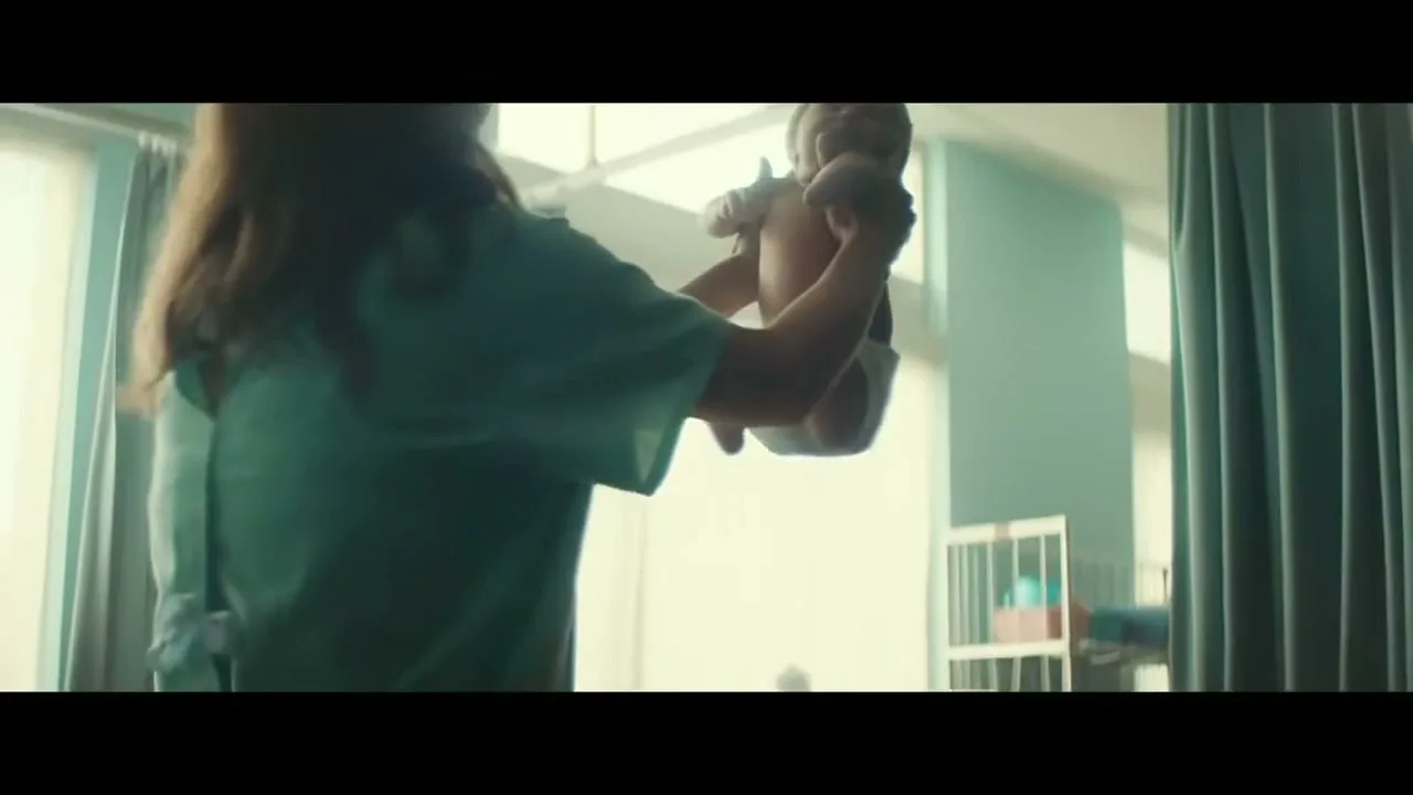 A nurse in teal scrubs holds a newborn baby up in the air, silhouetted against a bright window in a hospital nursery.