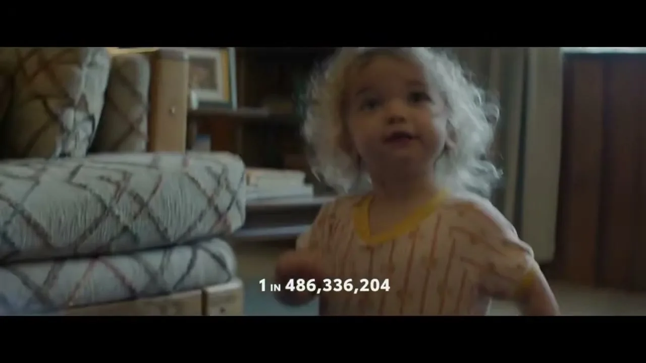 A toddler with curly blonde hair, wearing a striped onesie, takes her first steps in a living room, with text overlayed.
