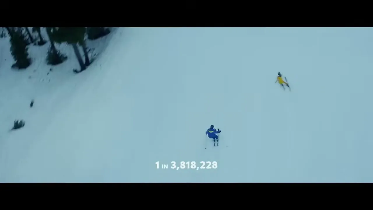 Two skiers, one in blue and one in yellow, ski down a vast, snow-covered mountain slope with trees visible on the left.