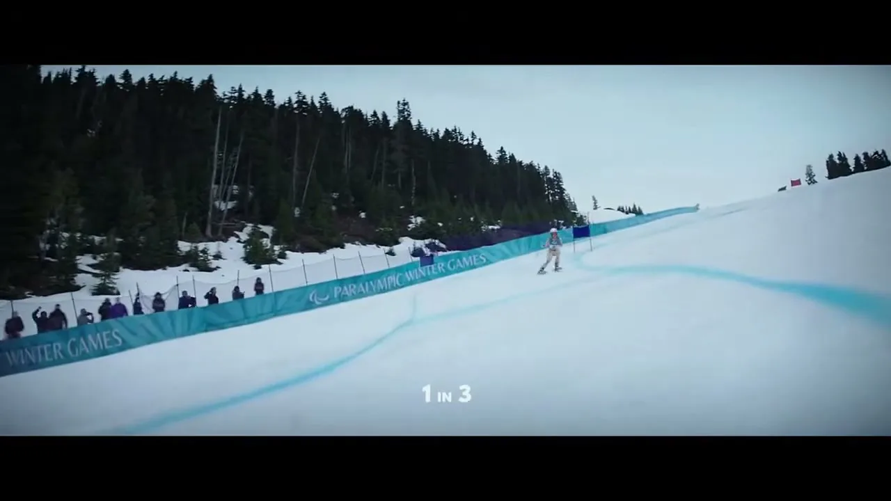 A para-alpine skier in a white and blue suit races down a snowy mountain slope, with spectators and pine trees in the background under a cloudy sky.