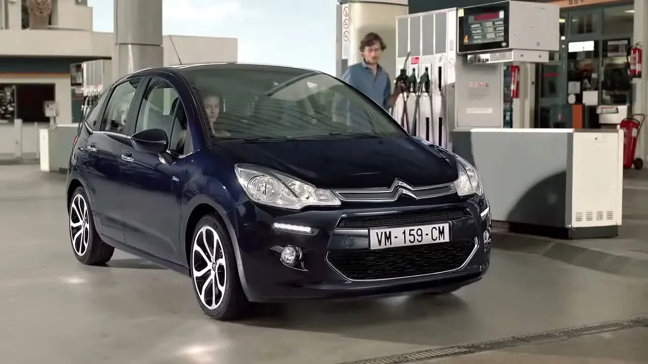 A dark blue Citroen C3 car is parked at a gas station, with a woman in the driver's seat and a man standing by the pump.