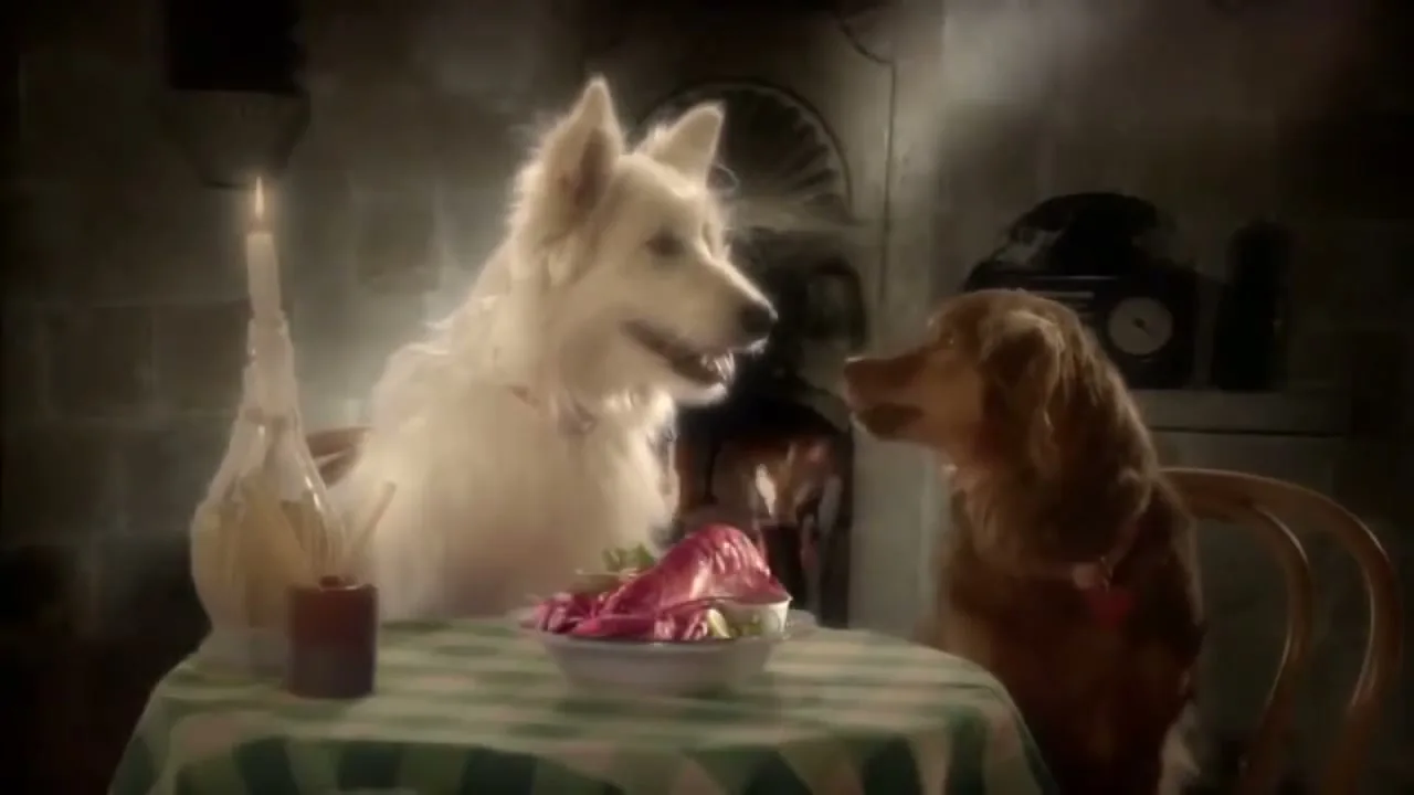 Two dogs, one white and one brown, sit at a small table with a checkered tablecloth, having a romantic dinner by candlelight in a dimly lit room.