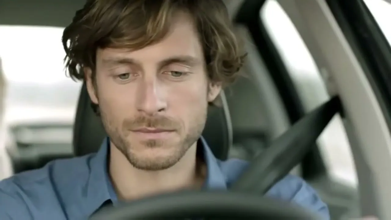A man with brown hair and a beard, wearing a blue shirt, looks down while driving a car.