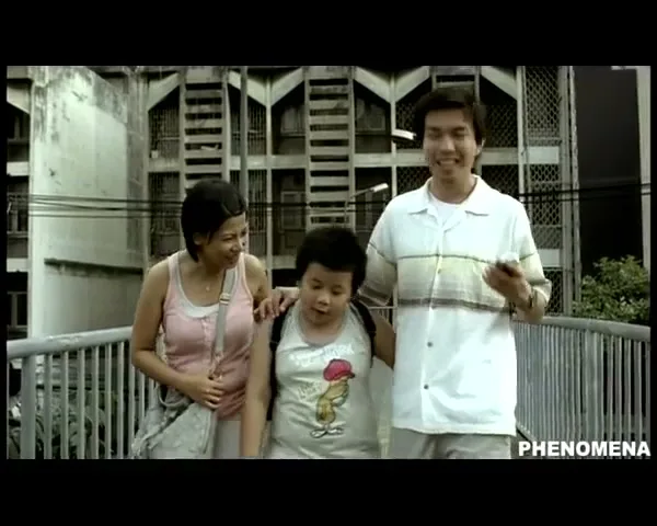 A family of three, a mother, father, and young son, walk across a pedestrian bridge, smiling and talking, with a building in the background.
