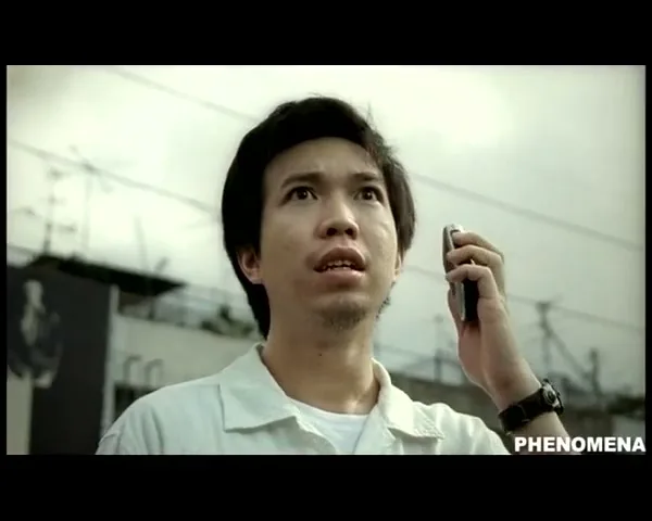 A man in his 30s, wearing a white shirt, holds a phone to his ear, looking up with a shocked expression on a city street.