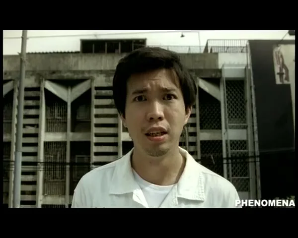 A young Asian man with short dark hair and a white shirt looks directly at the camera with a surprised expression in front of a building.