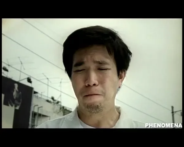 A man with dark hair and a light shirt cries with his eyes closed, standing outdoors with power lines and a billboard in the background.