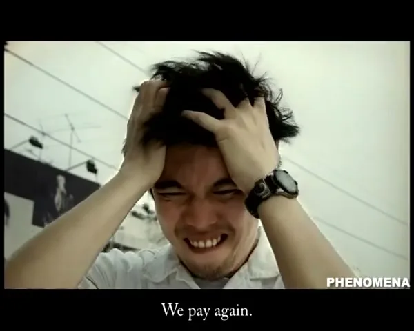 A man with dark hair and a goatee, wearing a white shirt, clutches his head in frustration, then looks surprised, outdoors during the day.