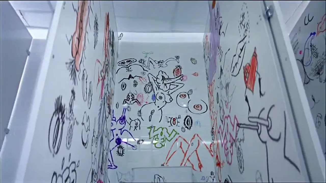 A wide shot of a public restroom stall covered in colorful, crude graffiti, with a toilet visible at the bottom.