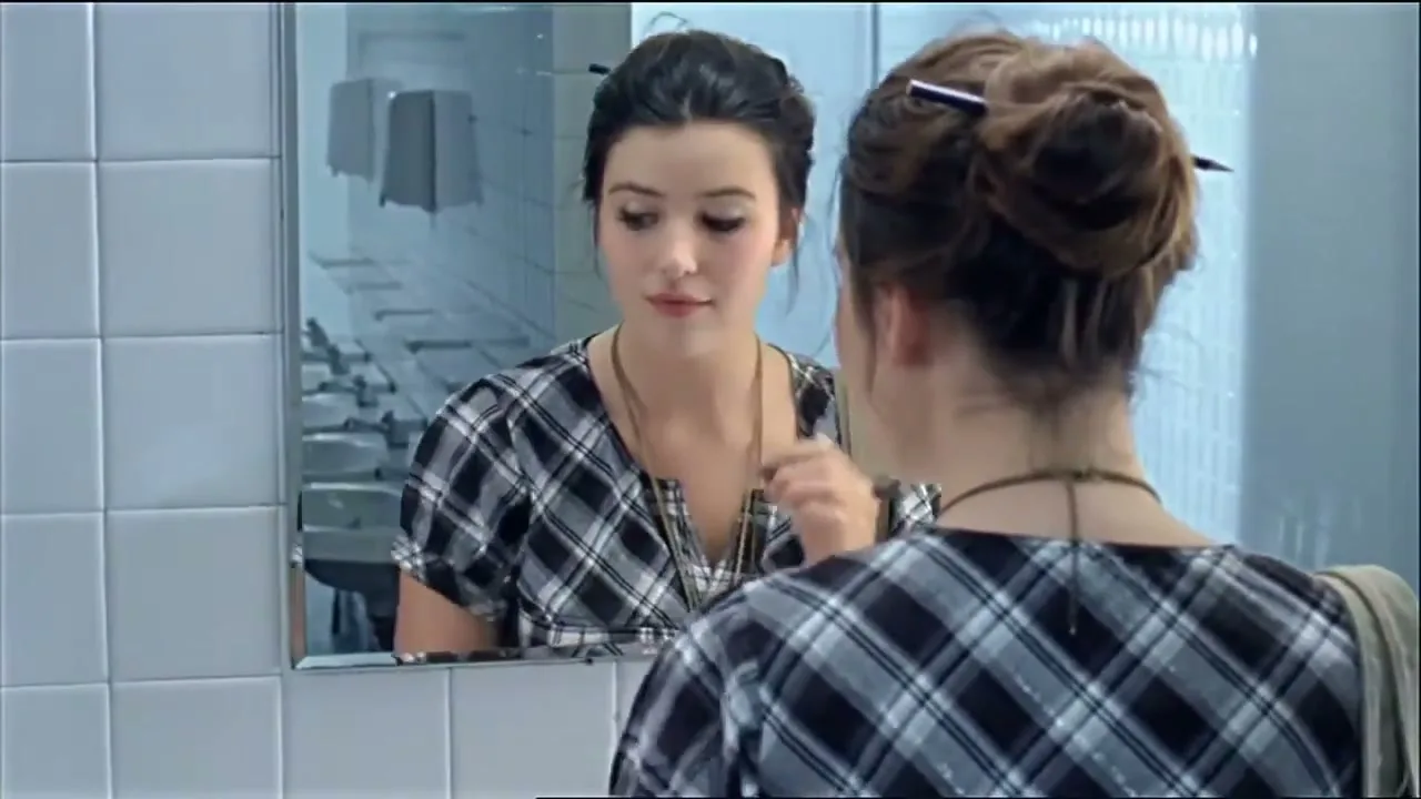 A young woman with dark hair in a plaid shirt applies eyeliner in a public restroom, then draws on the tiled wall with the eyeliner.