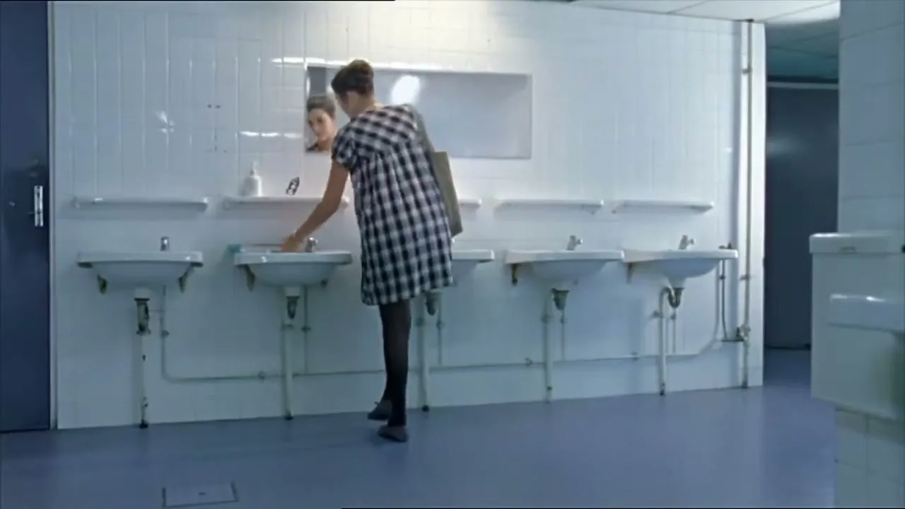 A young woman in a plaid dress and black tights washes her hands in a public restroom, then looks at her reflection in the mirror.