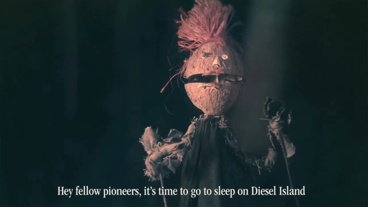 A stop-motion puppet made from a coconut and straw, with glowing eyes and a mustache, stands in a dark, smoky setting, speaking to the camera.
