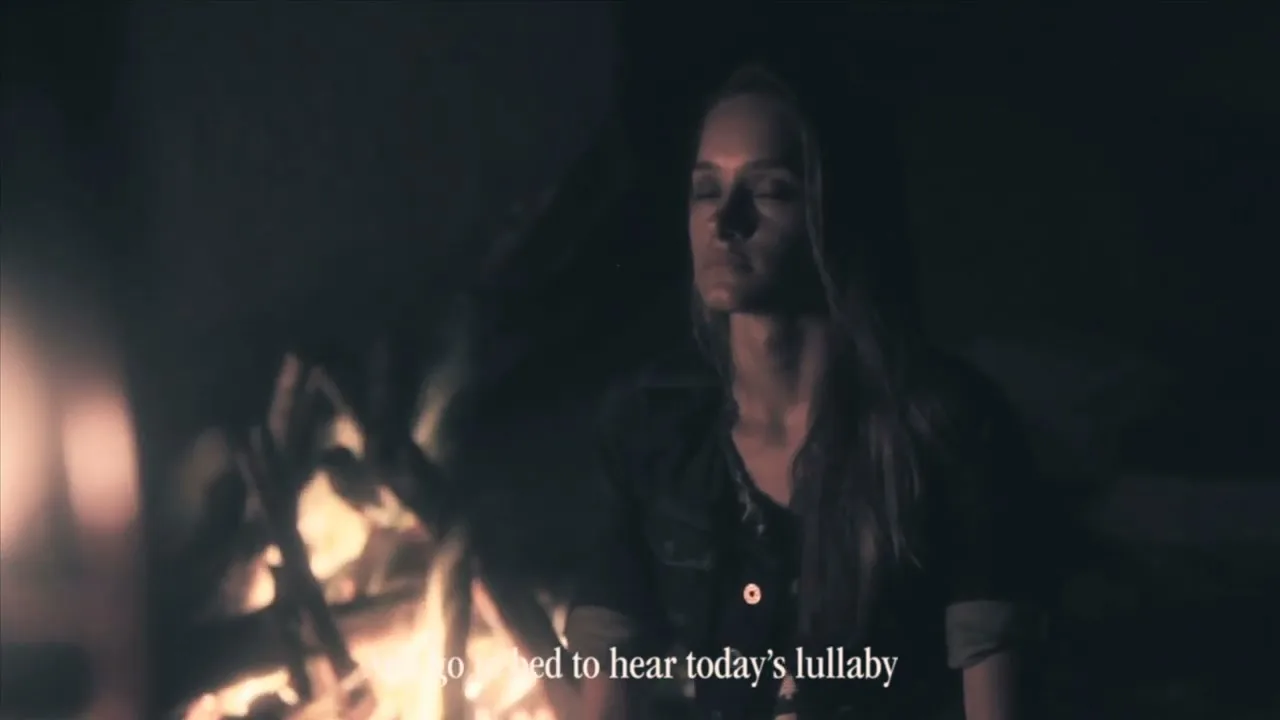 A young woman with long hair sits by a campfire at night, her eyes closed, in a dark, moody setting.