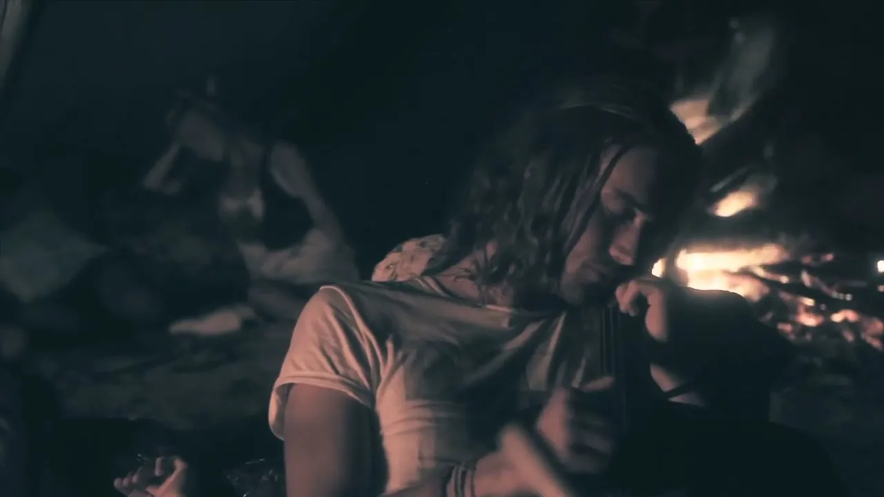 A young man with long hair, wearing a white t-shirt, plays a string instrument by a campfire at night, surrounded by friends.