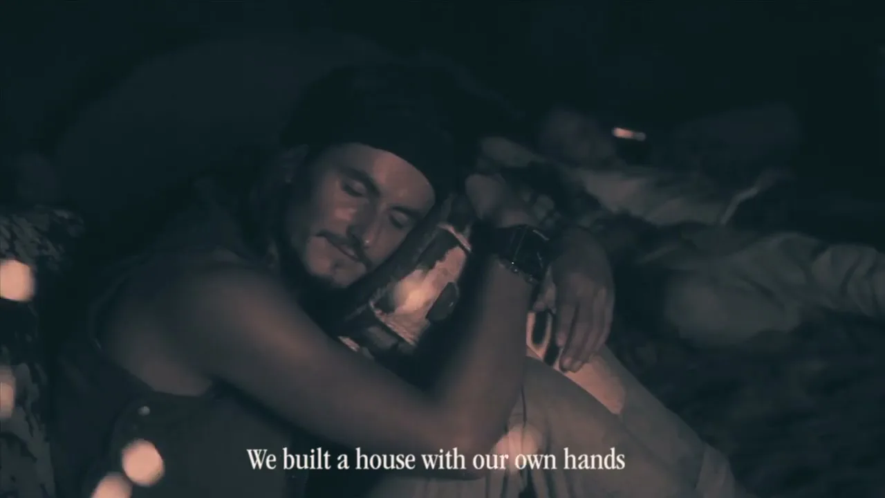 A man with a beard and a black bandana sleeps, hugging his knees, with other people sleeping in the background, in a dark, low-key setting.