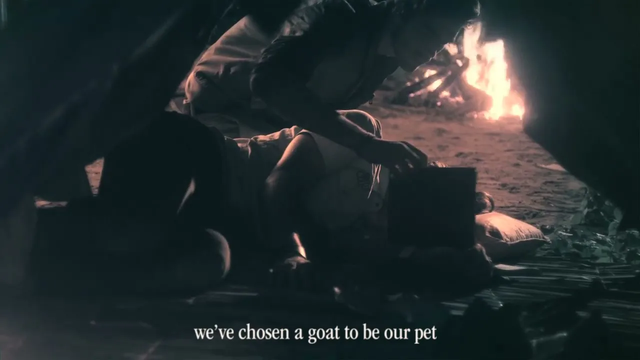 A young man lies on the ground, looking up at another person who is tending to him, next to a campfire at night.