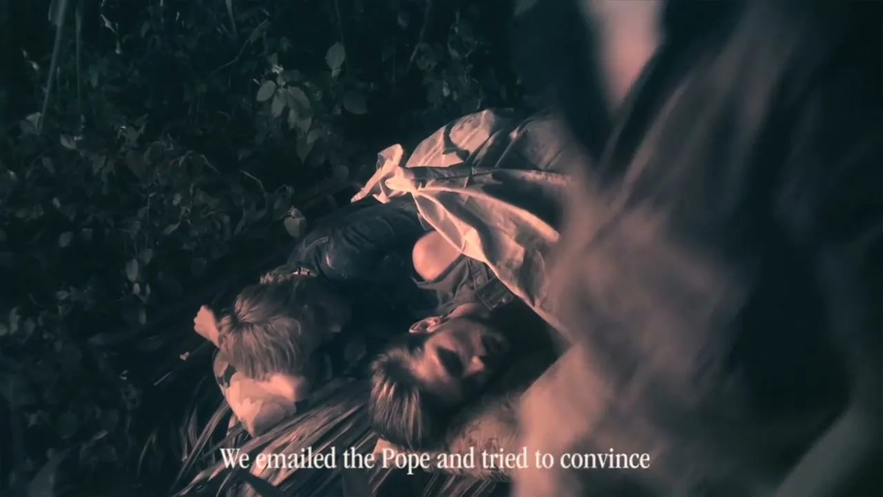 Two young men, one with blonde hair and one with brown hair, lie on the ground, covered in leaves and fabric, as another person leans over them in a dark, outdoor setting.