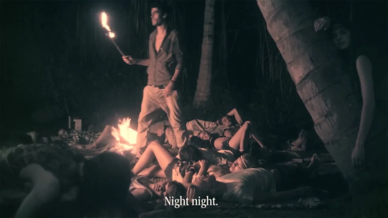 A man stands holding a torch over a group of people sleeping around a campfire on a tropical island at night.