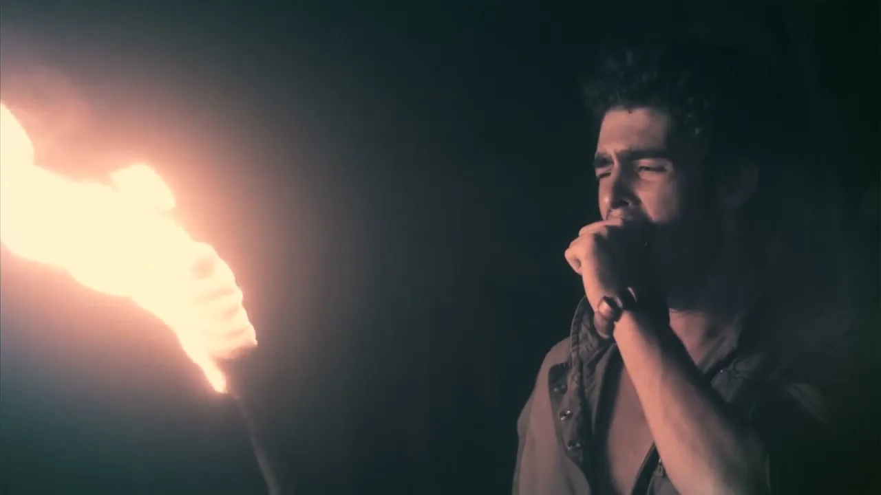 A man with dark hair, wearing an open shirt, holds a torch and looks distressed in a dark, indeterminate setting.