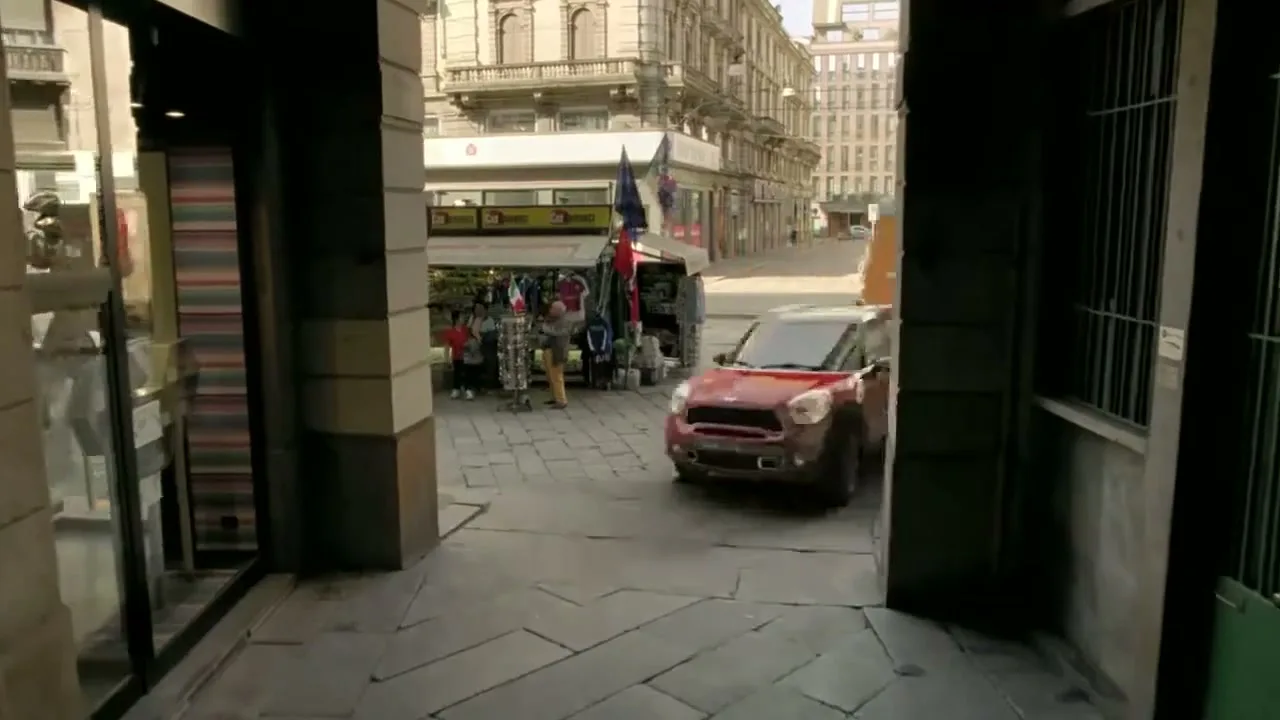 A red Mini Countryman drives quickly through a narrow, arched passageway in a European city, passing a street vendor's stall.