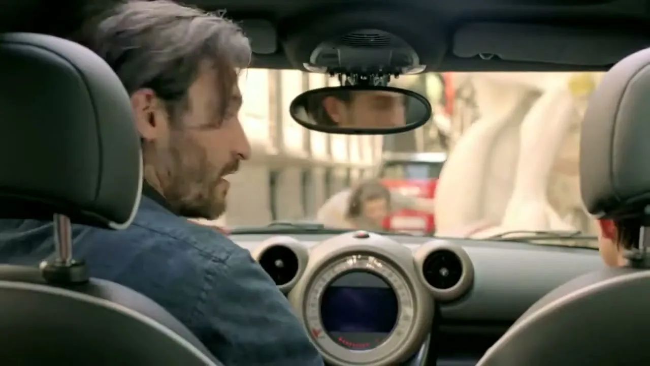 A man with a beard and denim shirt looks back from the driver's seat of a Mini Countryman, with a woman in the passenger seat, on a city street.