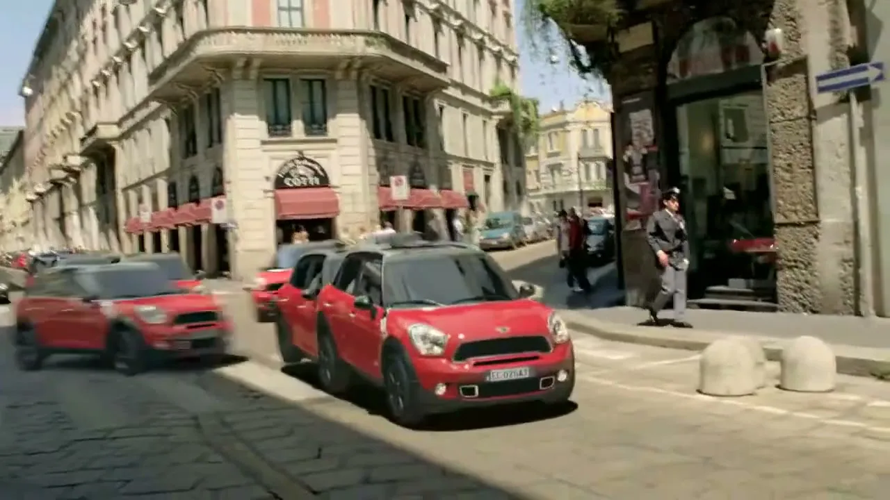 A line of red Mini Countryman cars drives down a sunny, narrow street in a European city, passing pedestrians and historic buildings.