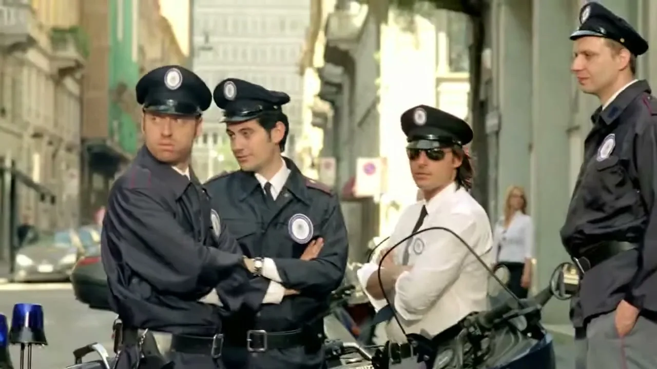 Four male police officers in uniform stand on a city street, two of them looking off-camera with confused expressions.