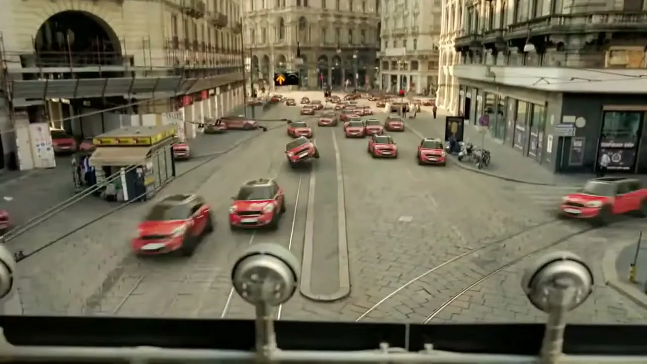 A wide shot from a tram shows many red Mini Countryman cars driving and swerving through a busy European city street during the day.