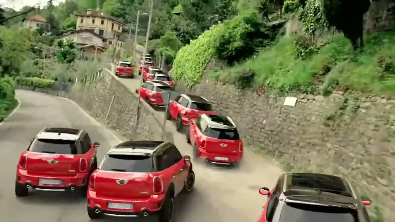 A long line of red Mini Countryman cars drives up a winding, narrow road in a lush, green, hilly landscape with buildings in the background.
