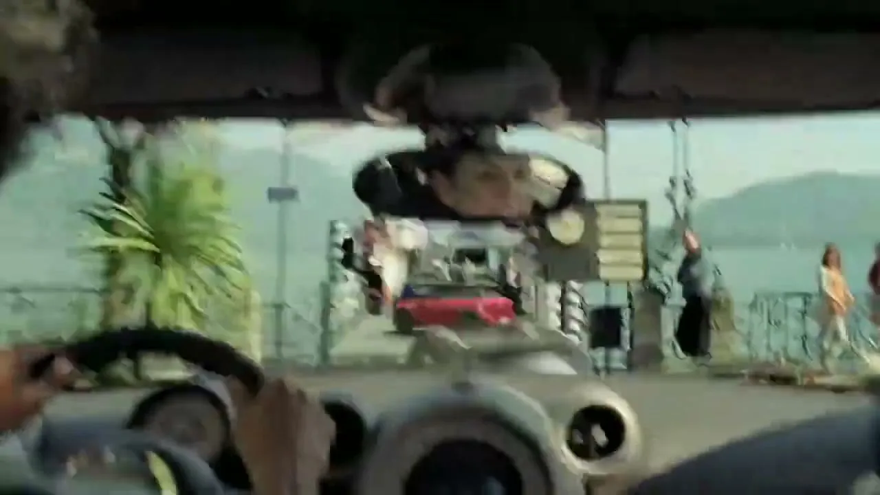 A driver's POV shot shows a red Mini Countryman backing up, with the driver's face reflected in the rearview mirror, at a lakeside ferry terminal.