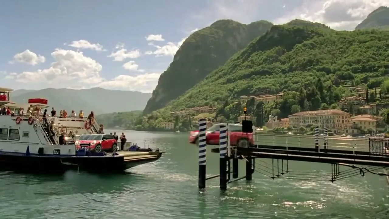 A red Mini Countryman drives off a ferry onto a pier on a sunny day, with a lake and mountains in the background.