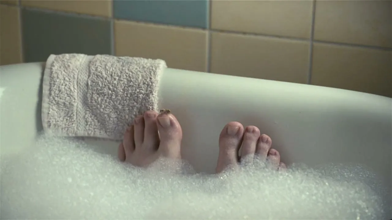 A person's feet are seen in a bubble bath, with a bee landing on one of their toes in a bathroom.