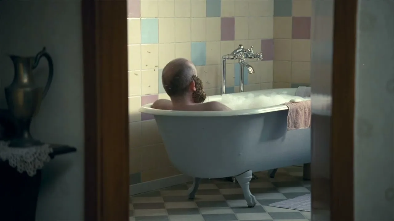 A bald man with a beard of bees relaxes in a bubble bath, seen through a doorway in a tiled bathroom.